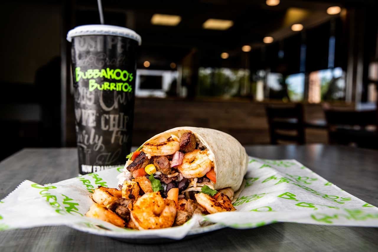 Bubbakoo's Burritos with steak and shrimp next to a cold fountain drink. Blurred restaurant in the background. 